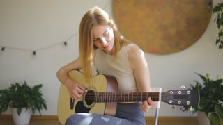 In a welllit, contemporary living room, a young woman with blonde hair showcases her musical skills and by playing an acoustic guitar